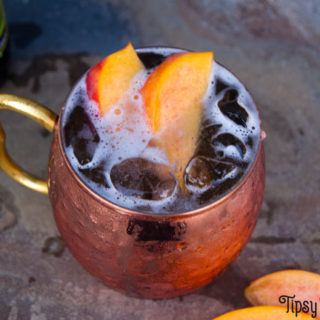 each Moscow mule in hammered copper cup with peach slice garnish and saranac ginger beer in the background