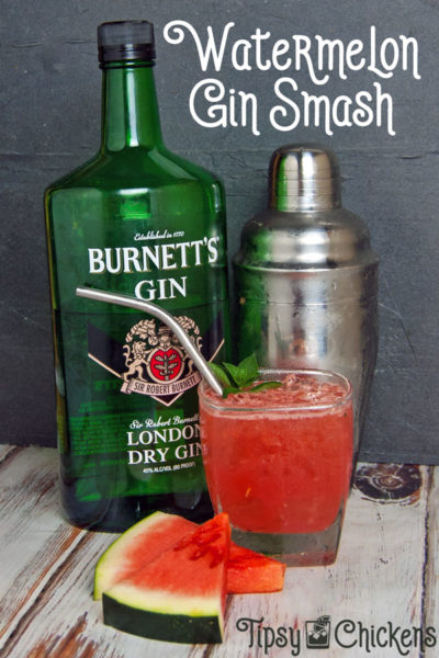 bottle of burnetts gin with a silver cocktail shaker and a square rocks glass filled with a watermelon gin smash garnished with mint and a metal straw with two slices of watermelon in the foreground