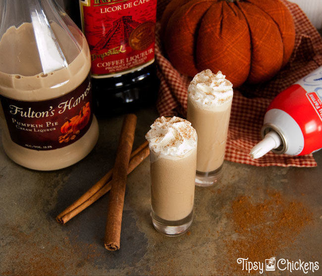 two tall shot glasses filled with pumpkin spice latte shots topped with whipped cream and a sprinkle of cinnamon on a mottled tile back ground with cinnamon sticks, a fabric pumpkin, Fulton's harvest pumpkin liqueur, kapali coffee liqueur and whipped cream in the background