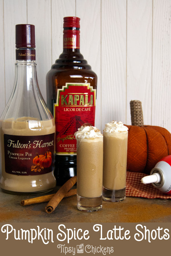 two tall shot glasses filled with pumpkin spice latte shots topped with whipped cream and a sprinkle of cinnamon on a mottled tile back ground with cinnamon sticks, a fabric pumpkin, Fulton's harvest pumpkin liqueur, kapali coffee liqueur and whipped cream in the background