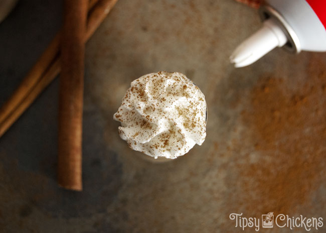 shot glass filled with pumpkin spice latte shots topped with whipped cream and a sprinkle of cinnamon on a mottled tile back ground with cinnamon sticks and whipped cream in the background