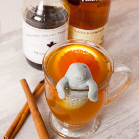 clear glass mug filled with Applejack and Ornage Hot toddy with Manatea tea strainer and bottles of maple syrup and applejack in the background on an aged white wooden surface wit h two crossed cinnamon sticks