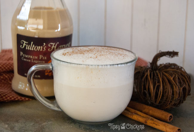 milk and pumpkin pie cream liqueur in a large clear coffee mug on a natural tile with a bottle of Fulton's Harvest Pumpkin Pie Cream Liqueur, cinnamon sticks and a wicker pumpkin