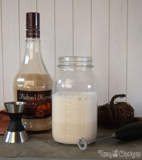 milk and pumpkin pie cream liqueur in a mason jar on a natural tile with a bottle of Fulton's Harvest Pumpkin Pie Cream Liqueur with a stainless steel jigger and an electric hand held frother