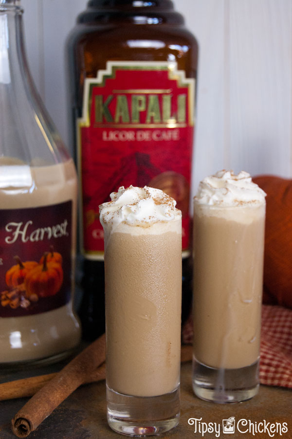 two tall shot glasses filled with pumpkin spice latte shots topped with whipped cream and a sprinkle of cinnamon on a mottled tile back ground with cinnamon sticks, a fabric pumpkin, Fulton's harvest pumpkin liqueur, kapali coffee liqueur and whipped cream in the background