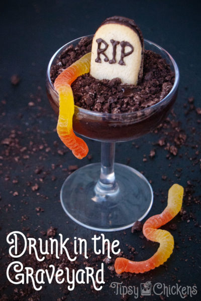 coupe glass filled with dark chocolate pudding made with alcohol with a tombstone cookie, crushed oreos and gummy worms