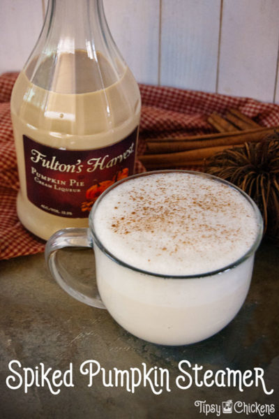 Large clear coffee mug filled with pumpkin steamer made with Fulton's Harvest Pumpkin Pie Liqueur on a mottles stone tile with a checkered cloth and a bottle of Fulton's Harvest Pumpkin Pie Liqueur