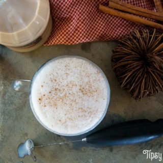 top view of milk and pumpkin pie cream liqueur in a large clear coffee mug on a natural tile with a bottle of Fulton's Harvest Pumpkin Pie Cream Liqueur, cinnamon sticks and a milk frother