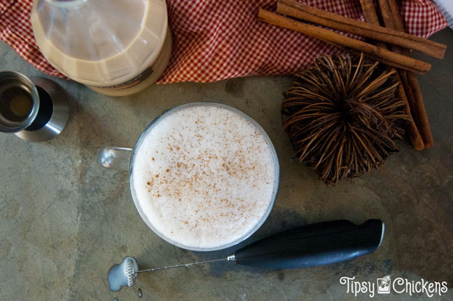 top view of milk and pumpkin pie cream liqueur in a large clear coffee mug on a natural tile with a bottle of Fulton's Harvest Pumpkin Pie Cream Liqueur, cinnamon sticks and a milk frother