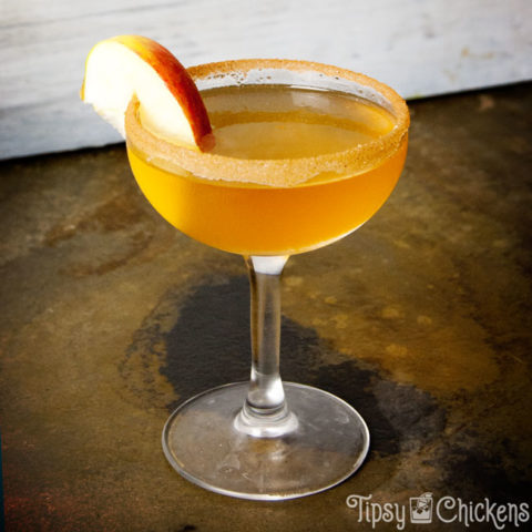 golden apple cider, cake vodka and bitters shaken together into an Apple Cider Donut cocktail served in a frosty coupe glass with a slice of apple on a cinnamon sugar coated rim