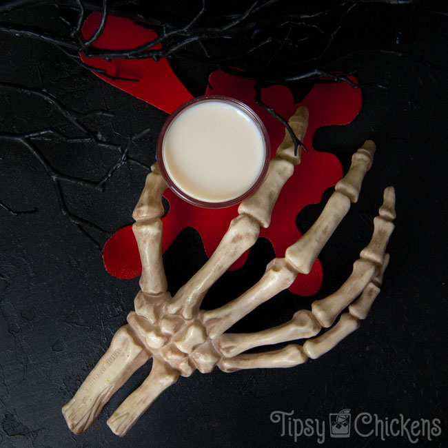 skeleton hand holding a shot glass filled with a Bone Daddy Halloween Shot over a red blood splatter coaster on a black background with spooky black branches