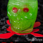 clear skull glass filled with crushed ice, rum, pineapple juice, Midori, blue Curacao with two red maraschino cherries for eyes on a black surface over a red blood splatter coaster