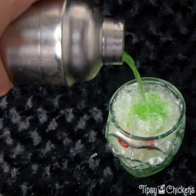 silver cocktail shaker pouring rum, pineapple juice, midori, blue Curacao into a clear skull glass filled with crushed ice with two maraschino for eyes on a black surface