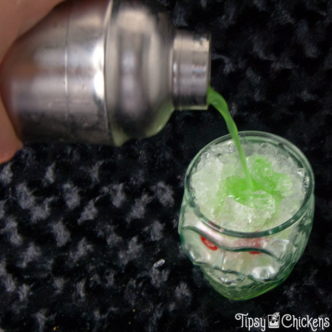  silver cocktail shaker pouring rum, pineapple juice, midori, blue Curacao into a clear skull glass filled with crushed ice with two maraschino for eyes on a black surface