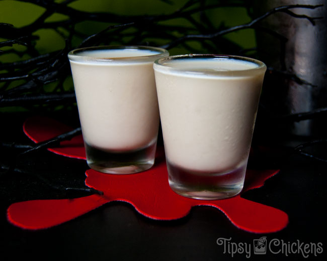 two shot glasses filled with Jack Skellington inspired Halloween shots sitting on a red blood splatter coaster with spooky trees and a green background