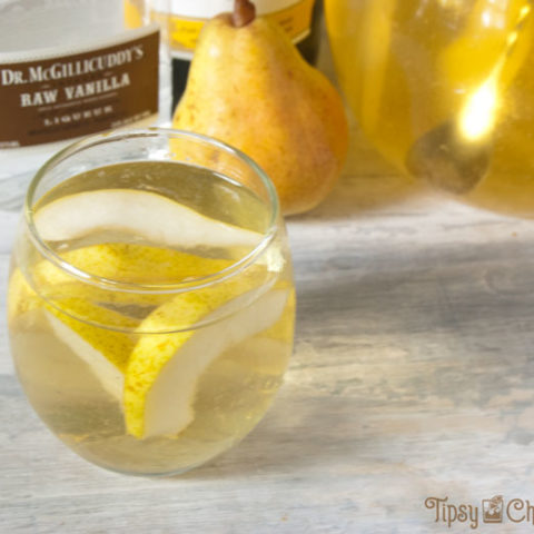 rounded glass and clear glass pitcher filled with sliced pears and white wine pear sangria with a bottle of chardonnay, a pear and a bottle of vanilla schnapps