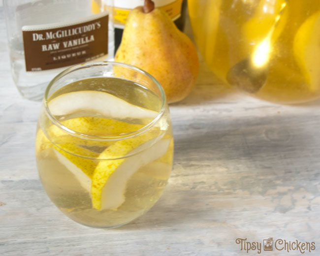 rounded glass and clear glass pitcher filled with sliced pears and white wine pear sangria with a bottle of chardonnay, a pear and a bottle of vanilla schnapps