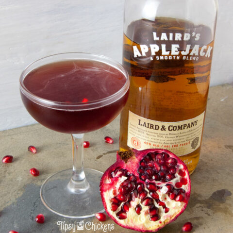 applejack, pomegranate cocktail in a coupe glass with a bottle of apple jack and half a pomegranate on a natural stone background