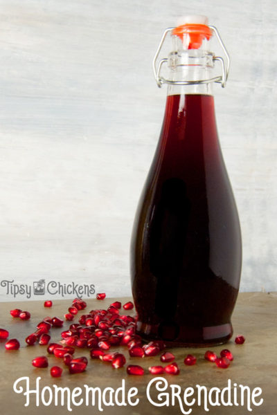homemade grenadine in a curvy glass bottle on a green stone tile with pomegranate seeds and a white background