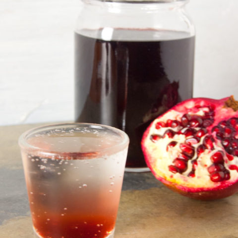 shot glass filled with clear soda with grenadine at the bottom with a mason jar full of grenadine in the background with half a pomegranate leaning on it