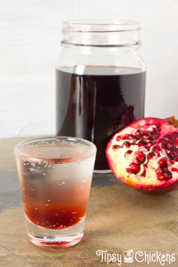 shot glass filled with clear soda with grenadine at the bottom with a mason jar full of grenadine in the background with half a pomegranate leaning on it 