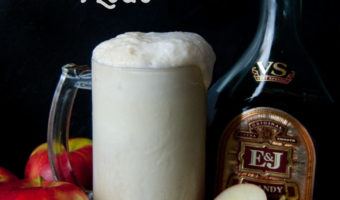 tall glass filled with vanilla ice cream, brandy, apple cider and ginger beer with a bottle of brandy and apples in the background and a single cut in half apple in front against a black background