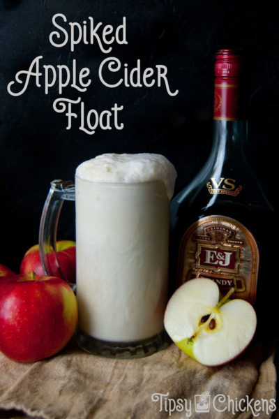 tall glass filled with vanilla ice cream, brandy, apple cider and ginger beer with a bottle of brandy and apples in the background and a single cut in half apple in front against a black background