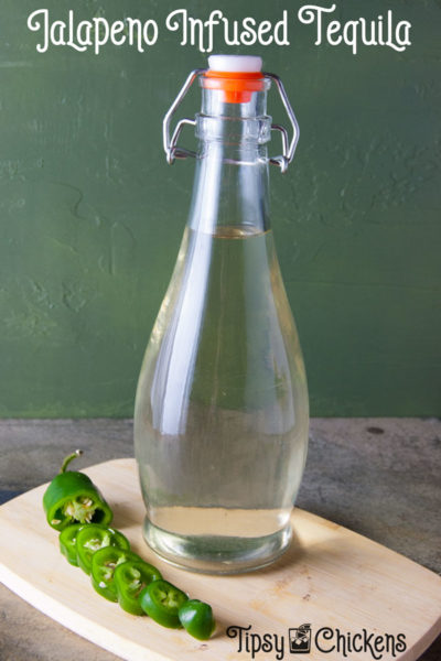 clear bottle filled with clear but slightly greenish tequila infised with jalapeno peppers sitting on a cutting board with a slided jalapno pepper to the left