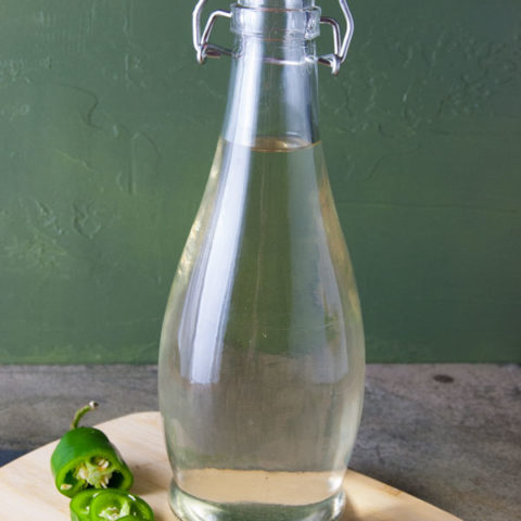 clear bottle filled with clear but slightly greenish tequila infised with jalapeno peppers sitting on a cutting board with a slided jalapno pepper to the left