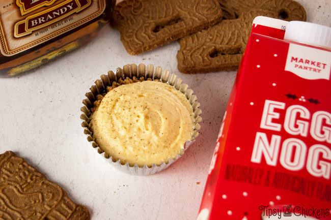 brandy spiked aggnog cheesecake in a metallic silver wrapper with Speculoos cookies, eggnog and brandy in the background