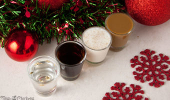 four shot glasses lined up on a white tile filled with different Christmas shots with red glitter snowflakes, red Christmas ornaments and a metallic red and green Christmas garland in the background