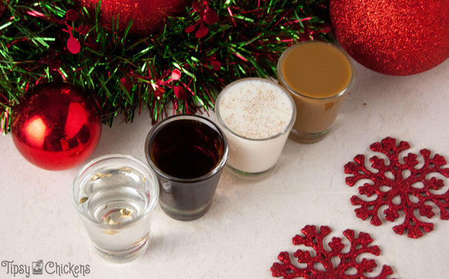 four shot glasses lined up on a white tile filled with different Christmas shots with red glitter snowflakes, red Christmas ornaments and a metallic red and green Christmas garland in the background 