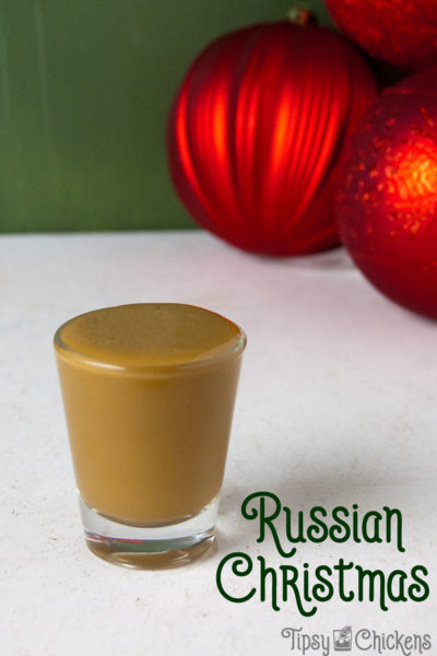 single shot glass filled with eggnog, coffee liqueur and vodka on a white tile surface with a green background and red Christmas balls