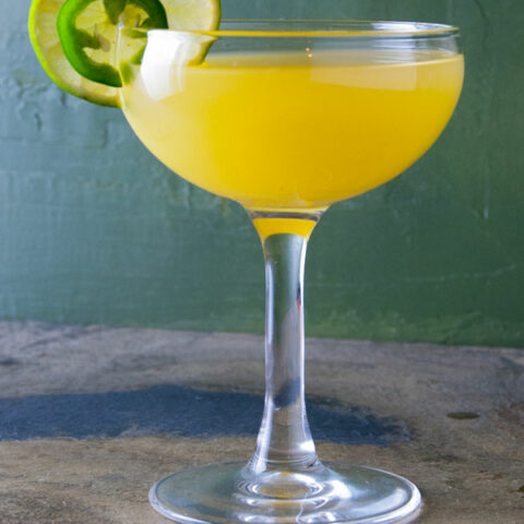 cocktail made with jalapeno tequila, pineapple juice and lime juice with a line wheel and jalapeno garnish against a green background