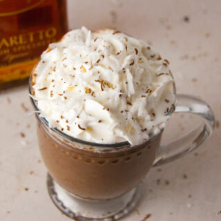 hot chocolate in a glass mug made with coconut milk, bakers chocolate, amaretto and coconut syrup topped with whipped cream and chocolate shavings with amaretto bottle in the background