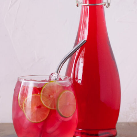 bottle of cranberry and lime infused vodka in a glass bottle with a clear glass cup filled with ice, lime slices and cranberry lime vodka soda with a metal straw