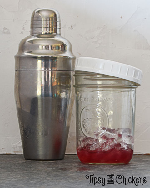 three piece cocktail shaker next to a mason jar with a white screw top lid filled with ice and a red liquid