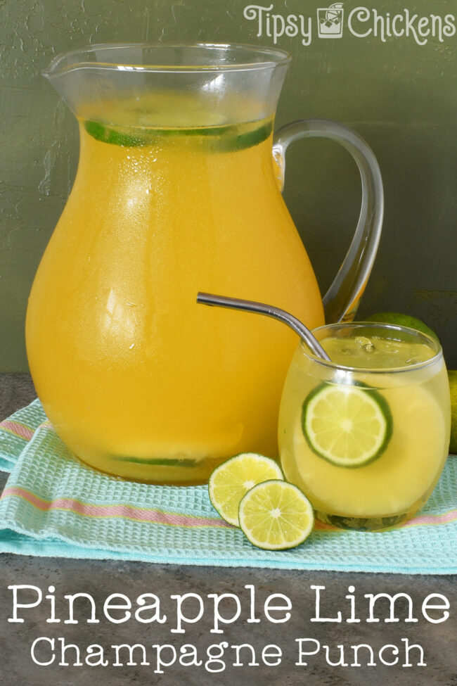 glass pitcher filled with pineapple juice, lime juice, ginger brandy, champagne and seltzer to make pineapple champagne punch on a blue towel over a grey tile with a rounded glass filled with champagne punch, a pineapple slice and a lime slice with a metal straw