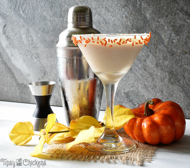 autumn pumpkin pie cocktail in a martini glass with a frosting and orange sprinkle rim with a cocktail shaker, jigger and fake pumpkin