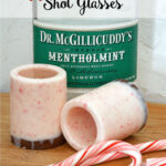 bottle of Dr. McGillicuddy's mentholmint behind two peppermint bark shot glasses made with almond bark and crushed candy canes on a cutting board with two candy canes